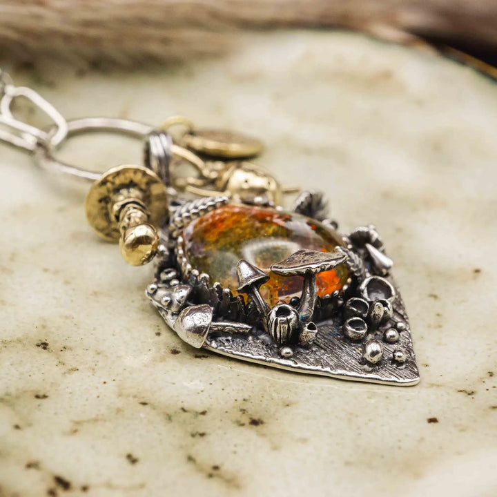 Statement Kette groß aus Silber 925. In den Anhänger eingefasst ist ein tropfenförmiger Lodolith, auch Gartenquarz genannt. Der Stein sieht aus wie eingeschlossenes grünes, gelbes und orangefarbenes Moos und ist umrandet von Pilzen, Blättern und Knospen. Ein echtes, handgefertigtes Statement Piece an Naturschmuck.
An dem Anhänger hängen außerdem 3 kleine Charms aus Messing: Ein Marienkäfer, ein Pilz und ein Blatt.