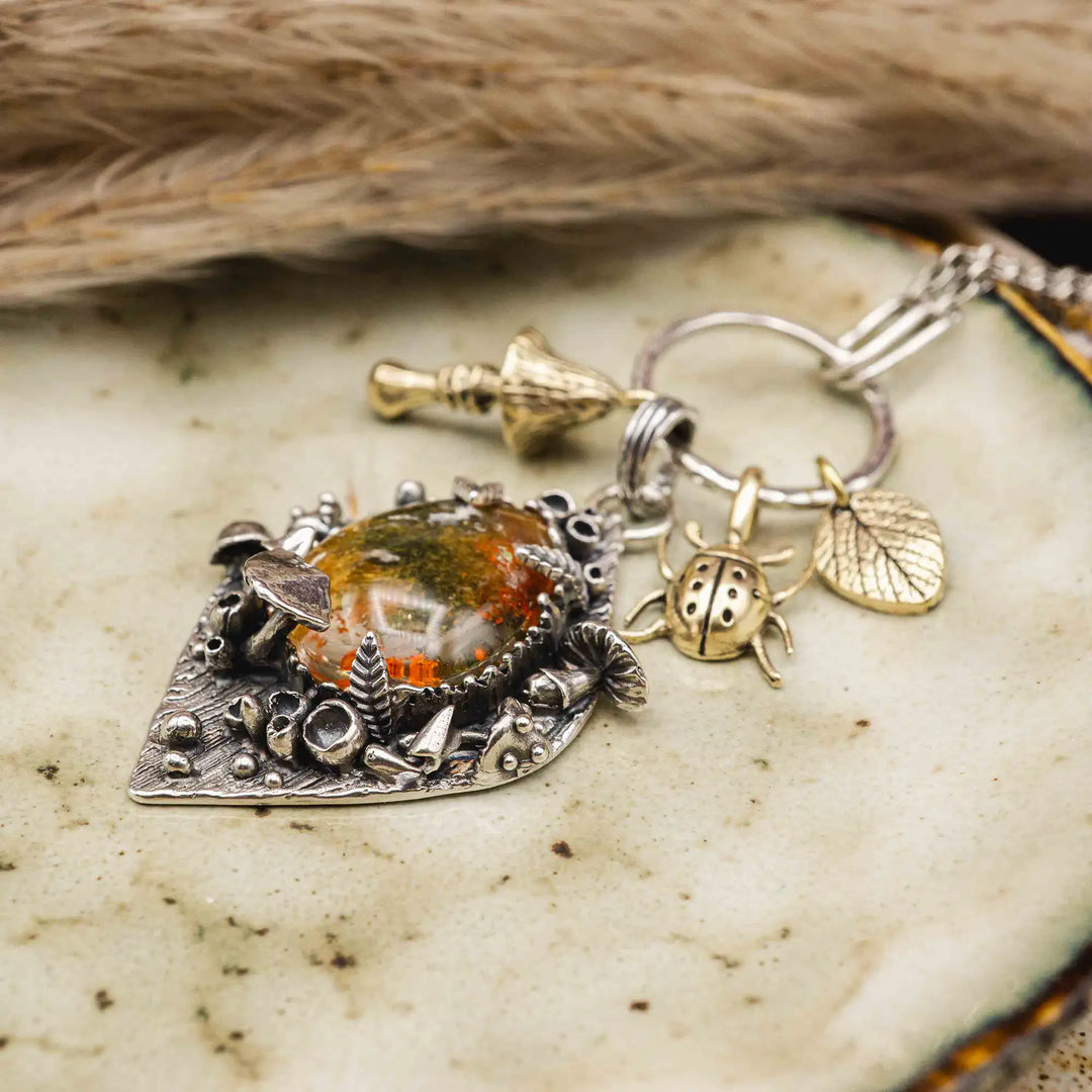 Statement Kette groß aus Silber 925. In den Anhänger eingefasst ist ein tropfenförmiger Lodolith, auch Gartenquarz genannt. Der Stein sieht aus wie eingeschlossenes grünes, gelbes und orangefarbenes Moos und ist umrandet von Pilzen, Blättern und Knospen. Ein echtes, handgefertigtes Statement Piece an Naturschmuck.
An dem Anhänger hängen außerdem 3 kleine Charms aus Messing: Ein Marienkäfer, ein Pilz und ein Blatt.
