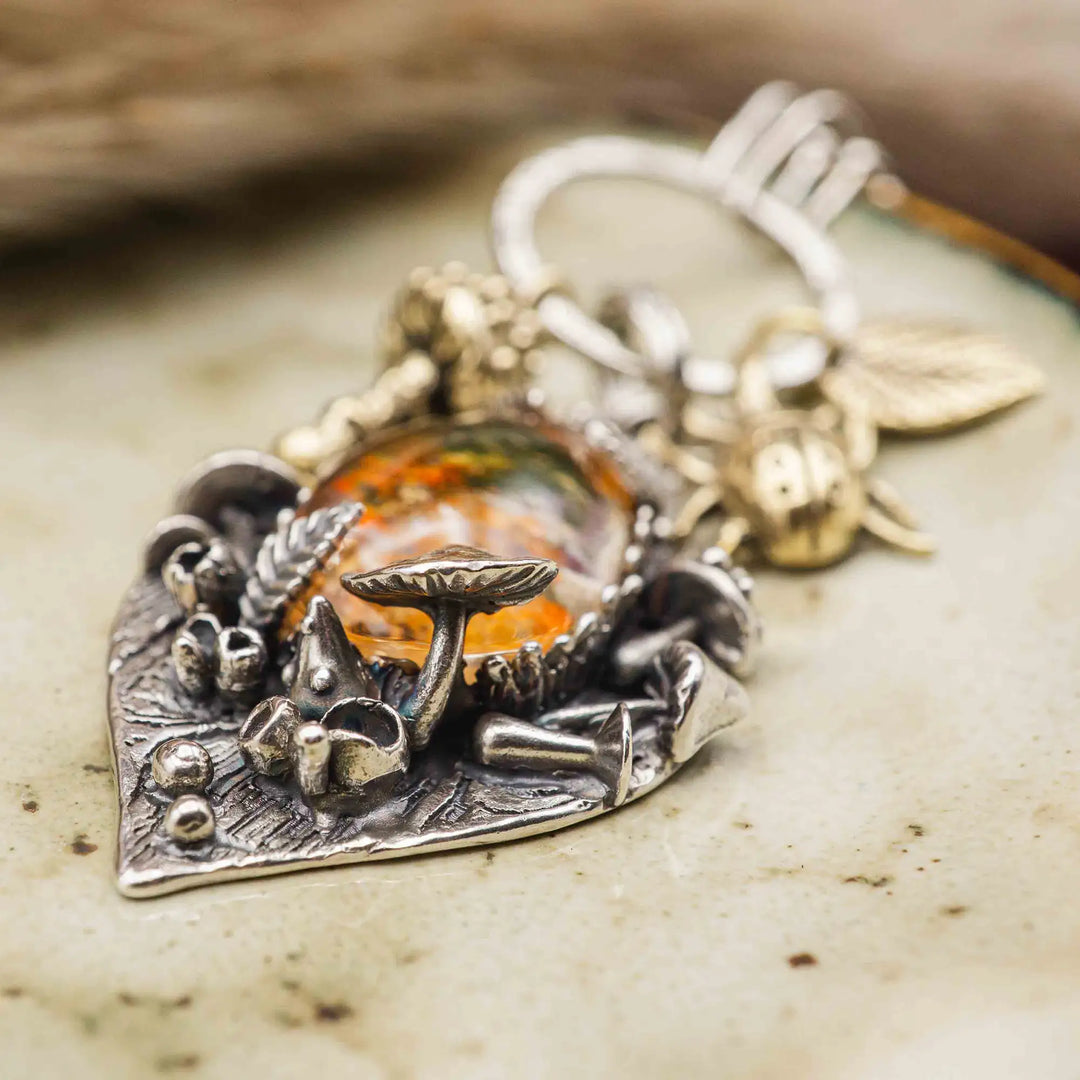 Statement Kette groß aus Silber 925. In den Anhänger eingefasst ist ein tropfenförmiger Lodolith, auch Gartenquarz genannt. Der Stein sieht aus wie eingeschlossenes grünes, gelbes und orangefarbenes Moos und ist umrandet von Pilzen, Blättern und Knospen. Ein echtes, handgefertigtes Statement Piece an Naturschmuck.
An dem Anhänger hängen außerdem 3 kleine Charms aus Messing: Ein Marienkäfer, ein Pilz und ein Blatt.